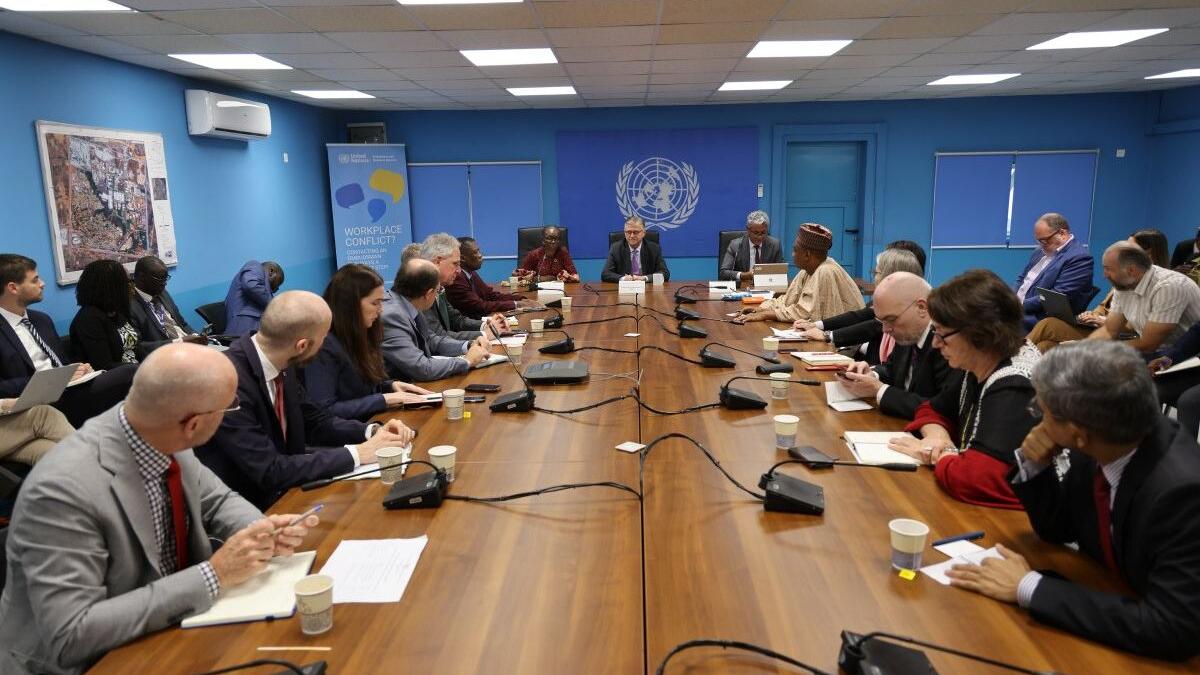 Meeting in a UN conference room with participants seated around a long wooden table, microphones and notebooks in front of them, and a UN emblem on the blue wall