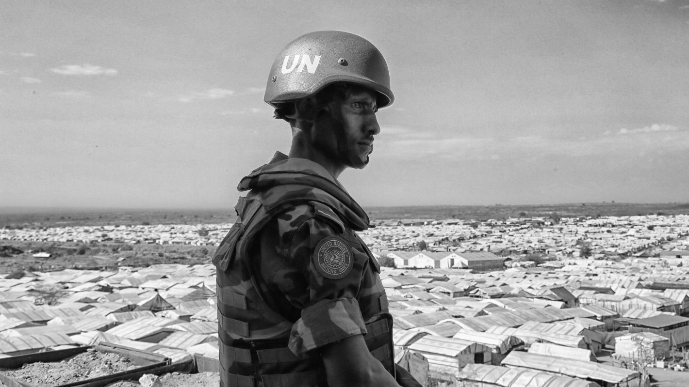 The UNMISS military component is embedded in coordination mechanisms at national, state and county levels that contribute towards a secure environment. It works to enhance trust between UNMISS and the South Sudanese government, makes efforts towards providing a stable and secure environment, thereby facilitating humanitarian access to populations in need. Additionally, the military engages the host government in capacity-building of its military institutions and reintegration of various armed groups. Photo by Nektarios Markogiannis/UNMISS. Black and white photo of a peacekeeeper surveilling buildings below.